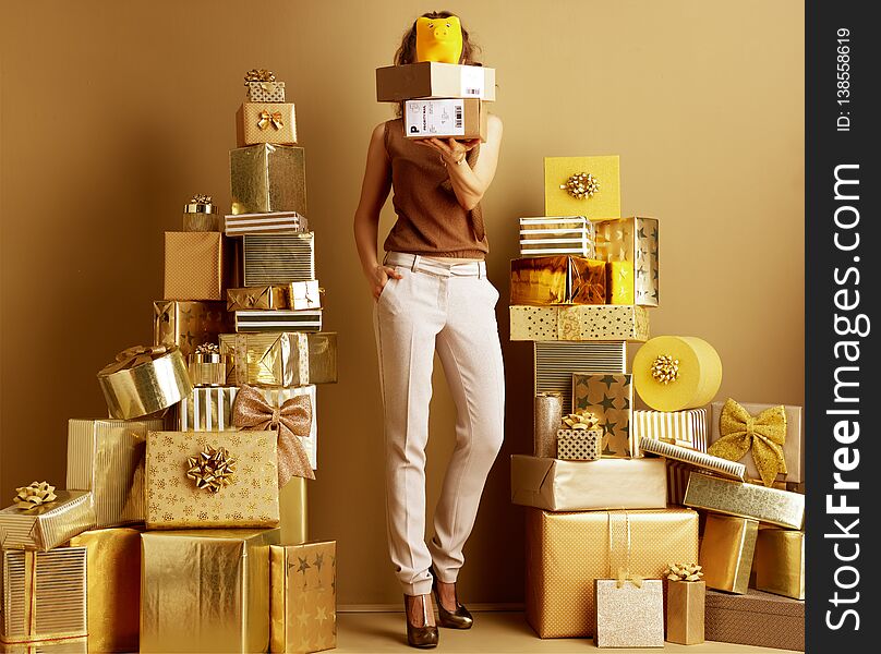 Stylish woman in gold beige pants and brown blouse among 2 piles of golden gifts in front of a plain wall holding parcels and yellow piggy bank in the front of face. Free holiday shipping. Stylish woman in gold beige pants and brown blouse among 2 piles of golden gifts in front of a plain wall holding parcels and yellow piggy bank in the front of face. Free holiday shipping