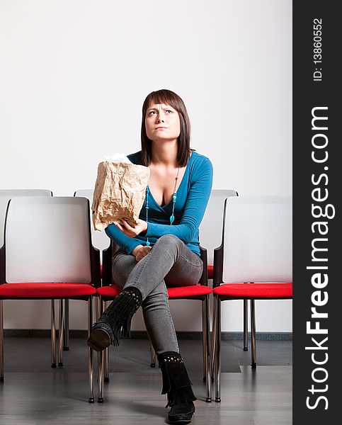 Worried young woman sitting with crossed legs in front cheer row, looking up, holding paper bag. Worried young woman sitting with crossed legs in front cheer row, looking up, holding paper bag