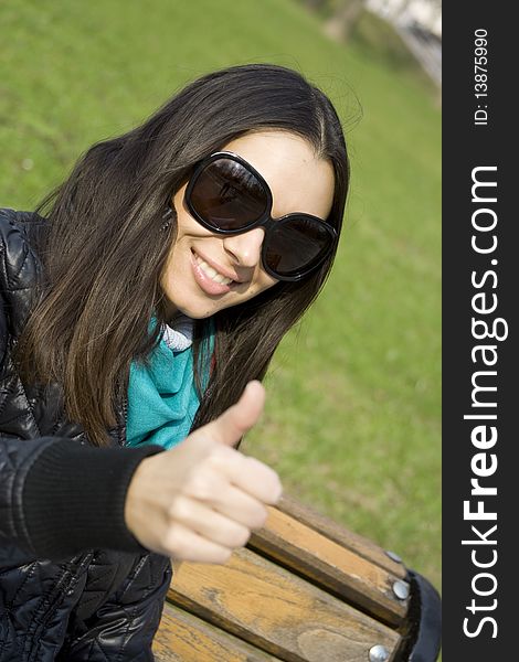 A beautiful young brunette girl in a park sitting on a bench smiling. Thumb sign OK. A beautiful young brunette girl in a park sitting on a bench smiling. Thumb sign OK