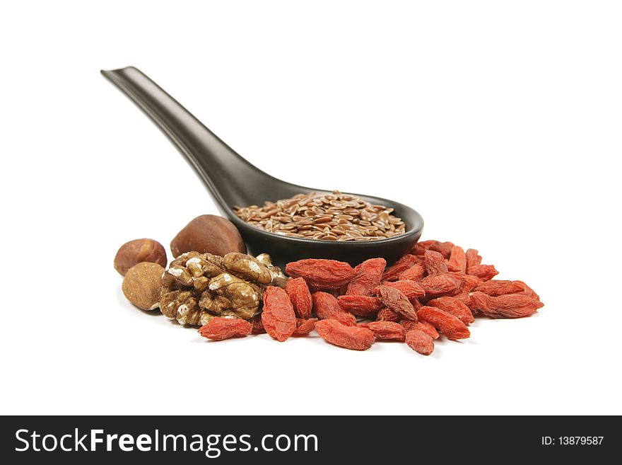 Brown linseed seeds on a small black spoon with mixed nuts and goji berries on a reflective white background. Brown linseed seeds on a small black spoon with mixed nuts and goji berries on a reflective white background