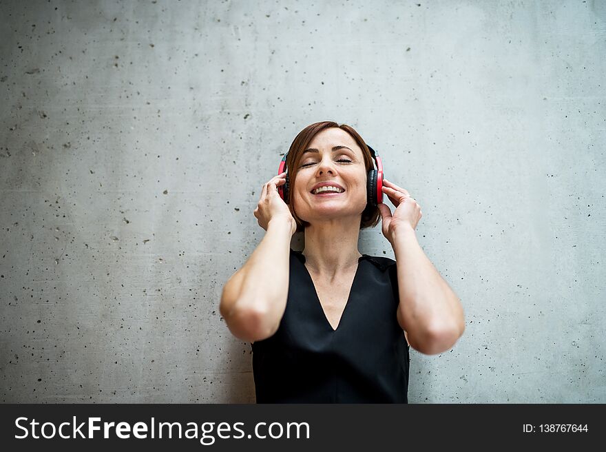 A portrait of young business woman with headphones standing against concrete wall in office and listening to music, eyes closed. A portrait of young business woman with headphones standing against concrete wall in office and listening to music, eyes closed.
