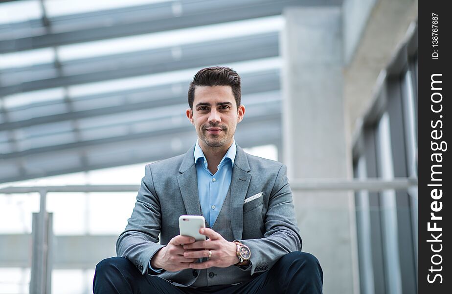 A portrait of young businessman with smartphone sitting in corridor outside office.