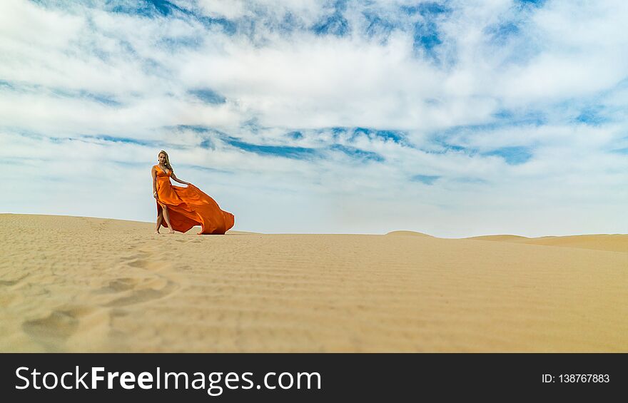 Girl walks on desert in dress fluttering in wind. Beautiful woman is walking, staing on sand or dune, touches, shows her