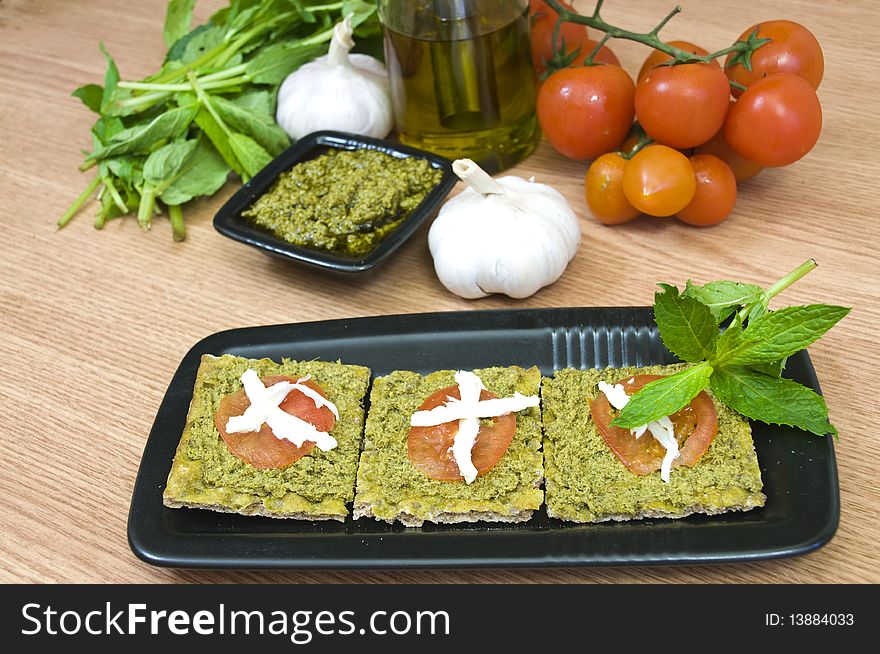 Pesto canapes and ingredients in the background; garlic, basil, tomatoes and olive oil