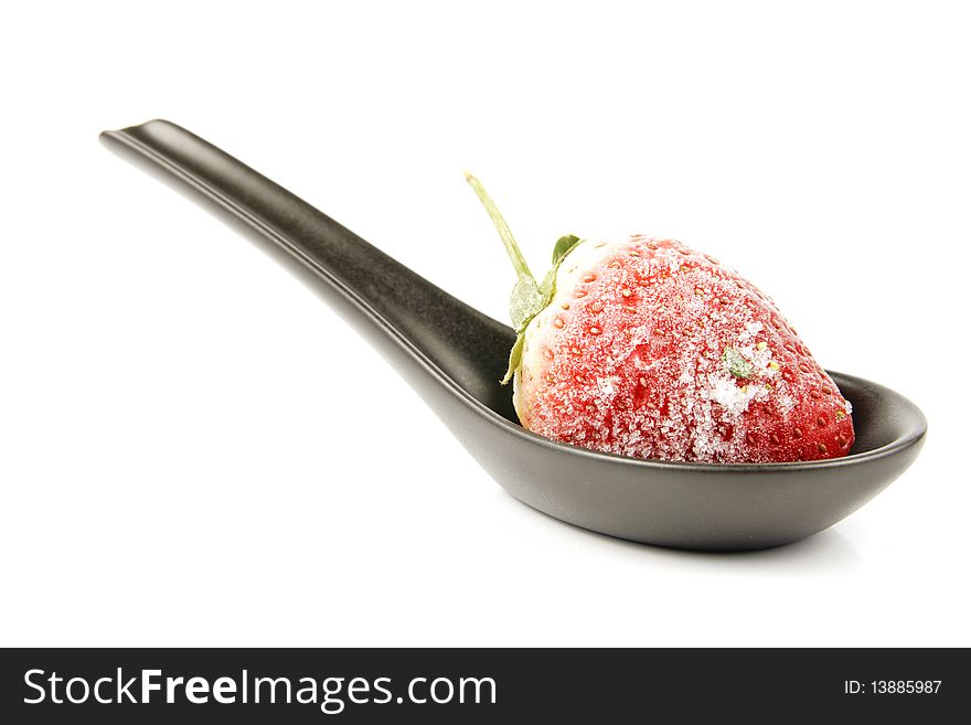 Red ripe frozen strawberry on a small black spoon with a reflective white background. Red ripe frozen strawberry on a small black spoon with a reflective white background