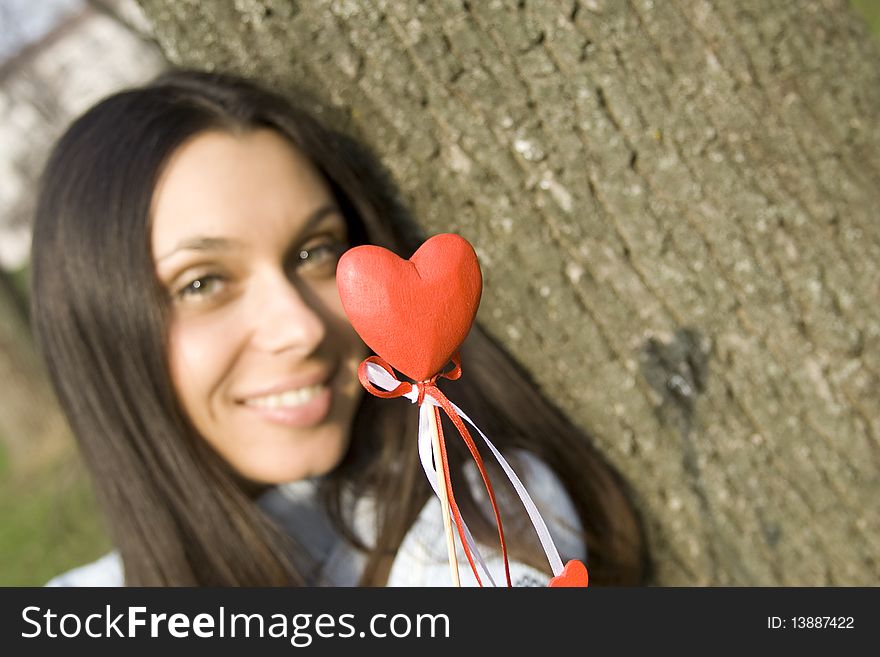 Beautiful girl in a park sitting on the grass near a tree in the hands of a red heart on the wooden stick. Beautiful girl in a park sitting on the grass near a tree in the hands of a red heart on the wooden stick