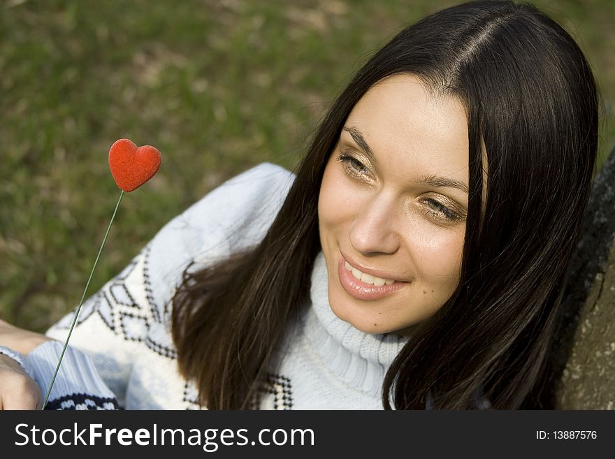 Beautiful girl in a park sitting on the grass near a tree in the hands of a red heart on the wooden stick. Beautiful girl in a park sitting on the grass near a tree in the hands of a red heart on the wooden stick