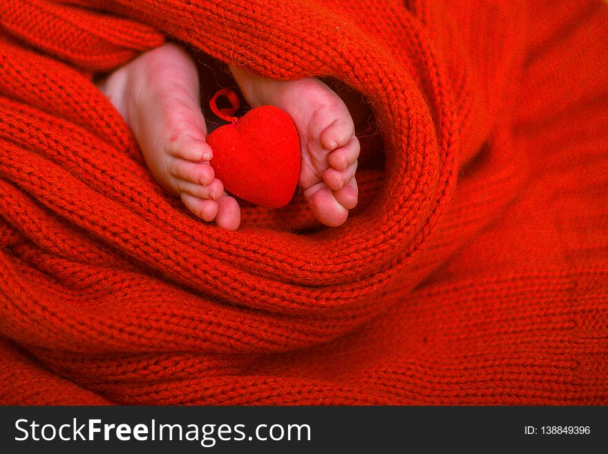 Red heart on baby legs. The legs of the newborn on a red background. A baby wrapped in a red blouse. Valentine`s Day