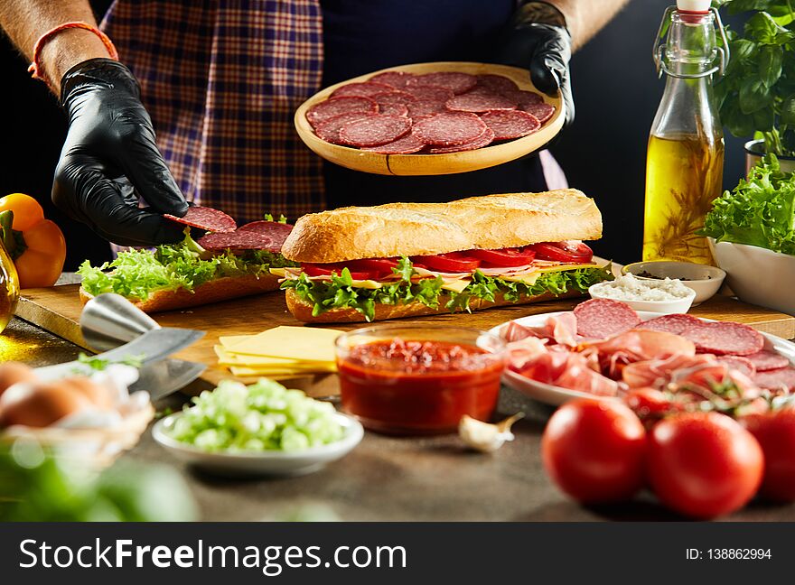 Takeaway chef preparing baguette sandwiches placing salami on the crusty bread with salad trimmings in a close up on his hands