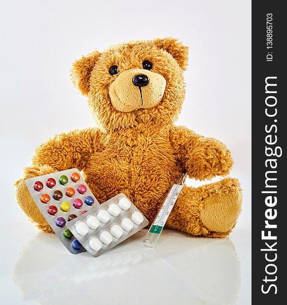 Toy bear with medicine, thermometer and colorful pills in blister pack, sitting on glossy surface against white background. Viewed from its front. Pediatrician office and children health concept