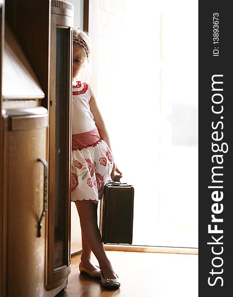 Pretty little girl being shy peeking out behind wooden furniture. blonde wearing headband and skirt holding a suitcase. Pretty little girl being shy peeking out behind wooden furniture. blonde wearing headband and skirt holding a suitcase.