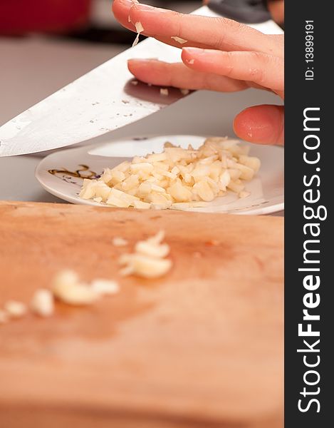 Cut garlic ready for cooking on a plate with cutting board in front.
Partially visible human hand removing the rest of the garlic from a knife.