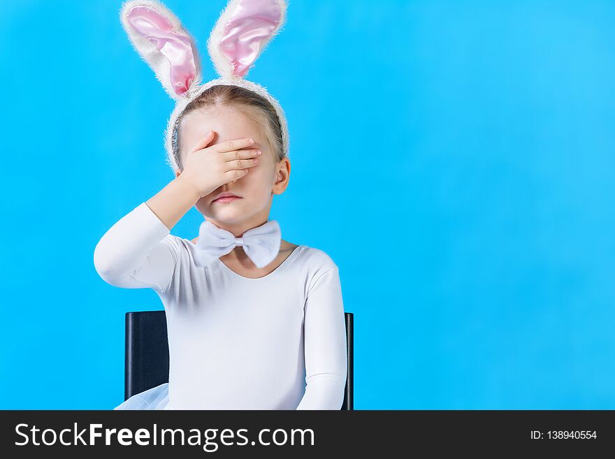 A little girl in a white rabbit costume closes her eyes with one hand. A child is sitting on a chair against a blue