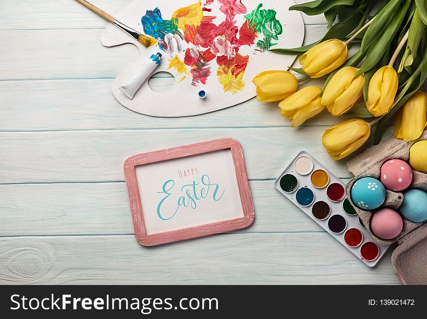 Colorful easter eggs, paint palette, brushes and yellow tulips on white wooden table. Top view with copy space.