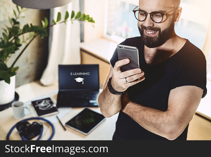 Portrait smiling handsome man reading text message, standing indoors. In background is laptop with inscription- e-learning on monitor.Businessman sends messages.Stylish brunette man uses mobile phone. Portrait smiling handsome man reading text message, standing indoors. In background is laptop with inscription- e-learning on monitor.Businessman sends messages.Stylish brunette man uses mobile phone