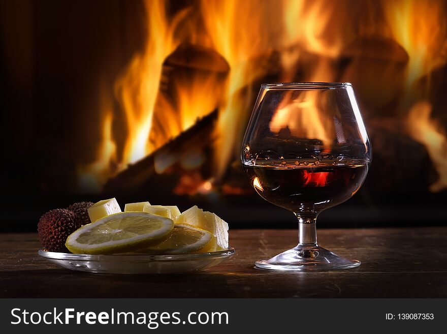 Still life of a glass of brandy and fruit with cheese on the background of a lit fireplace
