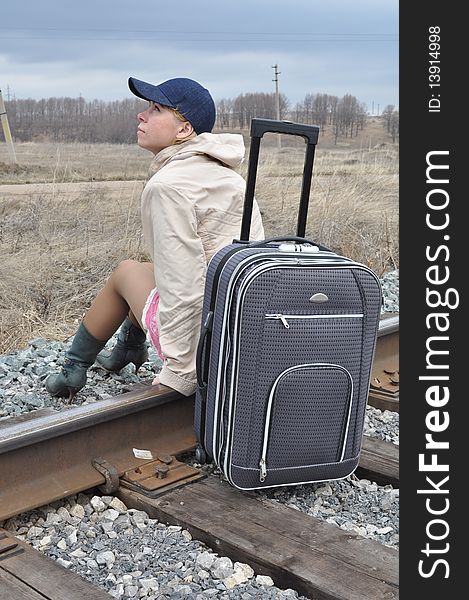 She is sitting with a bag on the rails, in a light jacket and a cap for baseball. She is sitting with a bag on the rails, in a light jacket and a cap for baseball