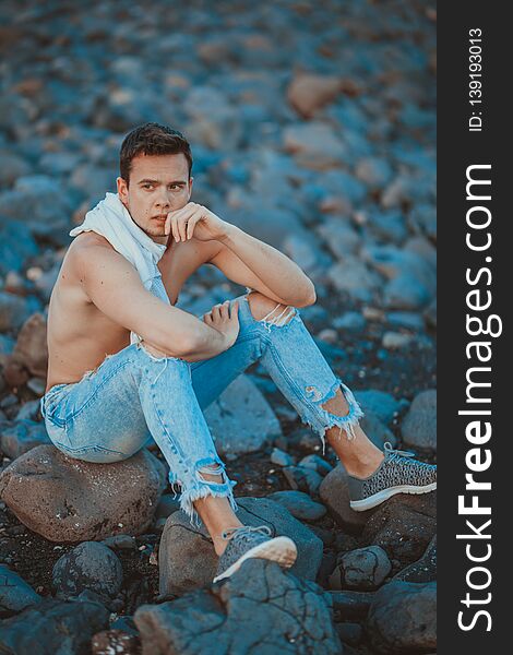 Photography of young man on the stone beach. Young man in casual outfit is sitting on the stone beach