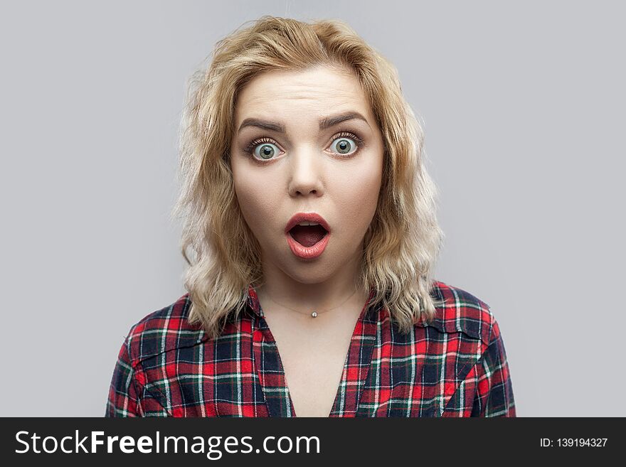 Closeup portrait of shocked beautiful blonde young woman in casual red checkered shirt standing with big eyes and looking at