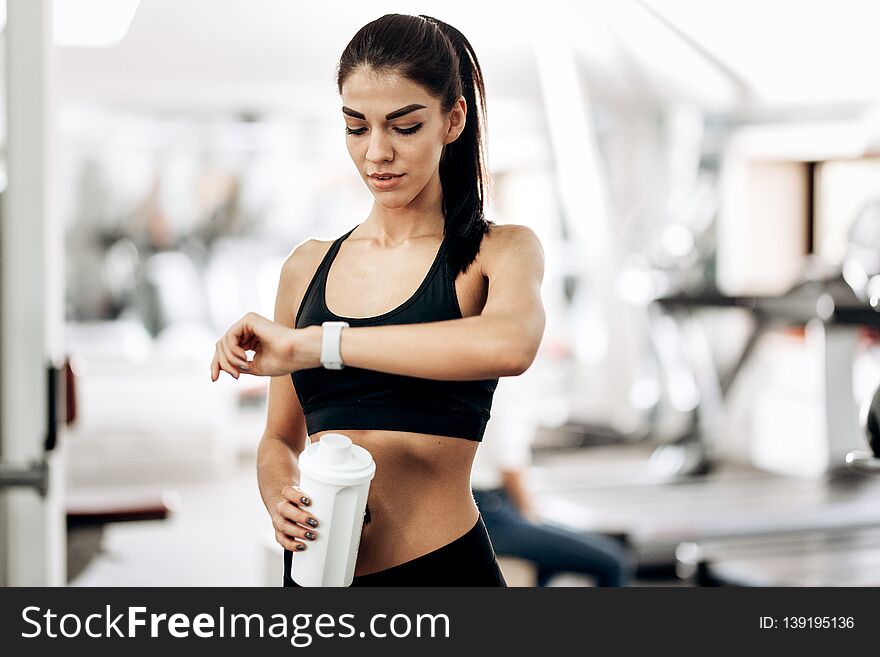 Beautiful slim girl dressed in black sportswear is standing in the gym and looking at her watch .