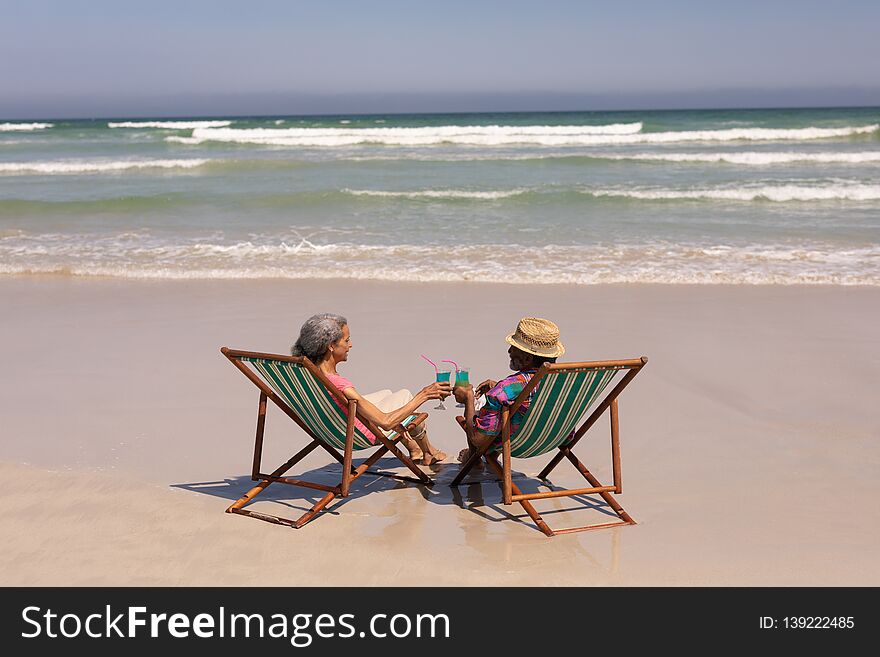 Rear view of senior couple relaxing on sun lounger and toasting cocktail glasses on beach in the sunshine