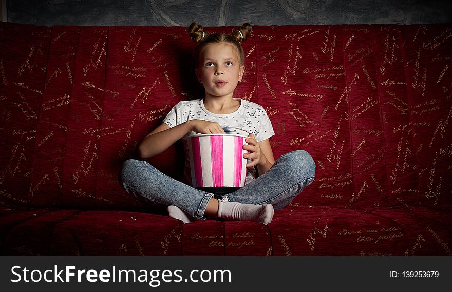 Children`s cinema: One schoolgirl girl watches a movie at home on a big red sofa and eats a popkort from a red bucket. Portrait.