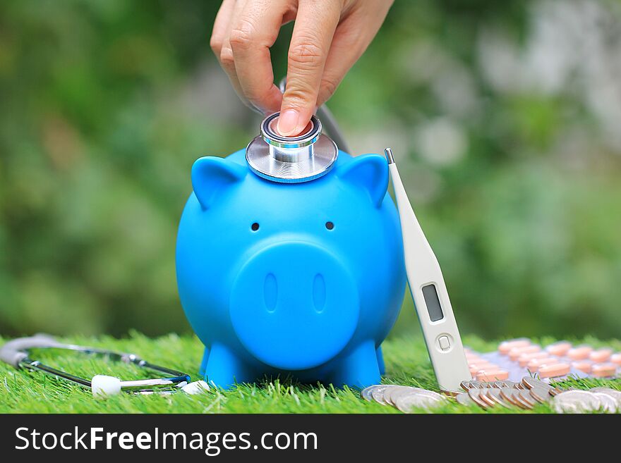 Woman using stethoscope to check blue piggy bank with thermometer and medicine on natural green background,Save money for Medical expenses and Health care concept