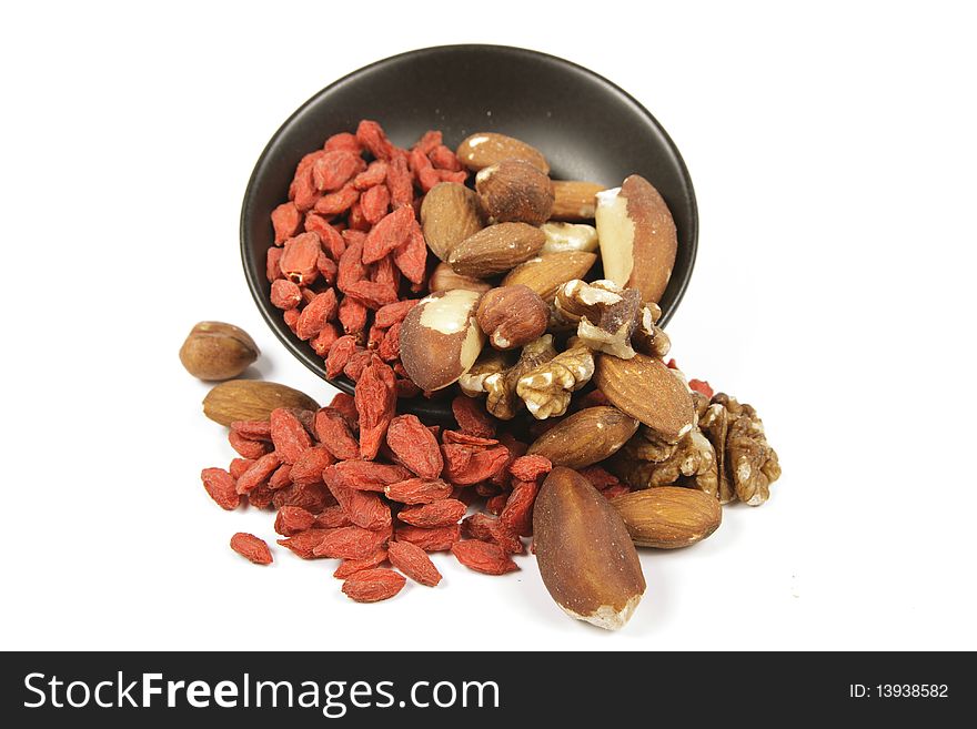 Red dry goji berries with mixed nuts spilling from a small black bowl on a reflective white background. Red dry goji berries with mixed nuts spilling from a small black bowl on a reflective white background