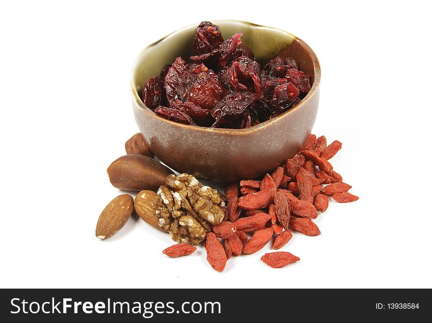 Red ripe dried cranberries in a small green and brown bowl with mixed nuts and goji berries on a reflective white background. Red ripe dried cranberries in a small green and brown bowl with mixed nuts and goji berries on a reflective white background