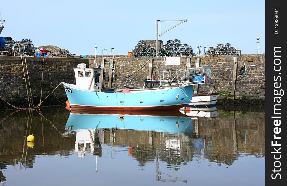 A small light blue fishing boat tied up in a harbour in west Wales. Lobster and crab pots are stacked on the quayside. The water is glassy and rippled. A small light blue fishing boat tied up in a harbour in west Wales. Lobster and crab pots are stacked on the quayside. The water is glassy and rippled.