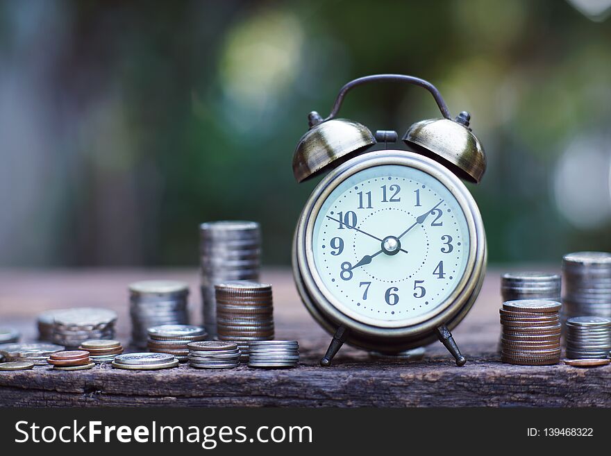 Vintage alarm clock and coin stacks on wooden table with blur green garden background, bright morning color tone, finance and