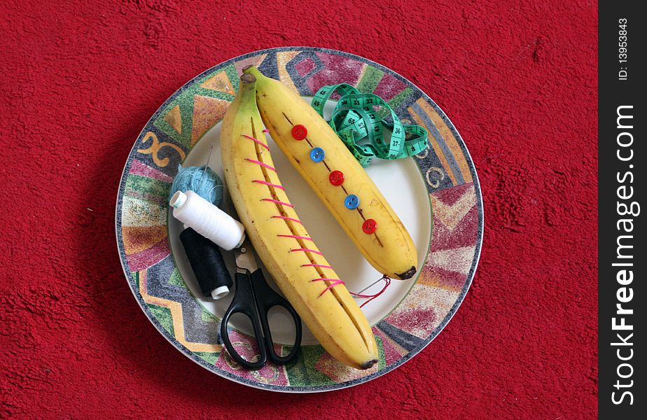 Two bananas on a plate, thread, scissors, a centimeter tape, buttons, a needle