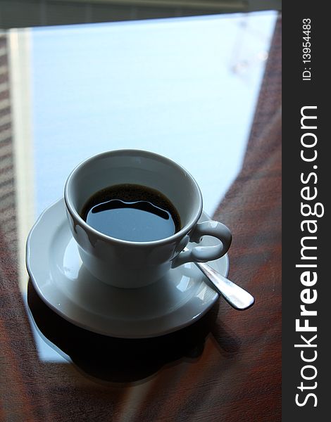 Close up view of a coffee  cup on a wood and crystal table under a window. Close up view of a coffee  cup on a wood and crystal table under a window