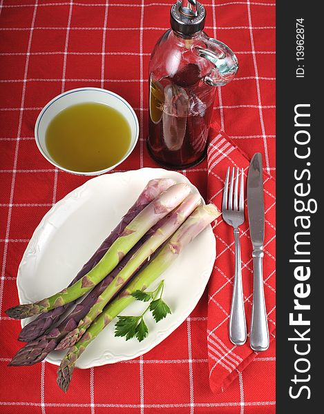 Bunch of fresh organic and home-grown asparagus on a white dish resting on a red table cloth  with a bottle of vinegar in the background next to a small bowl of olive oil. Folded napkin and silverware on top. Bunch of fresh organic and home-grown asparagus on a white dish resting on a red table cloth  with a bottle of vinegar in the background next to a small bowl of olive oil. Folded napkin and silverware on top.