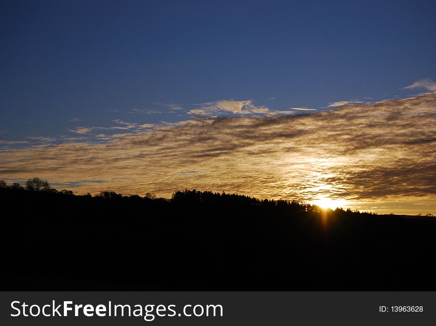 Sunset, with clouds against blue sky, above dark pine ridge