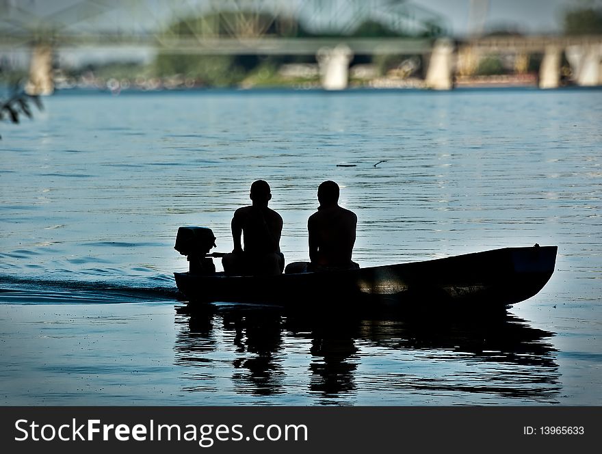A two man in a boat, afternoon light silhouettes, bridge in out on focus background