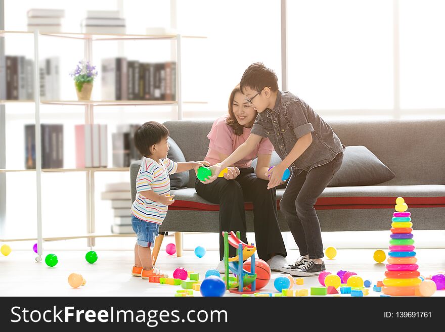 Asian family, happy time at home, young beautiful mother, taking care sons, little adorable toddler, standing, young boy wearing eyes glasses hold green ball, playing together in modern living room. Asian family, happy time at home, young beautiful mother, taking care sons, little adorable toddler, standing, young boy wearing eyes glasses hold green ball, playing together in modern living room