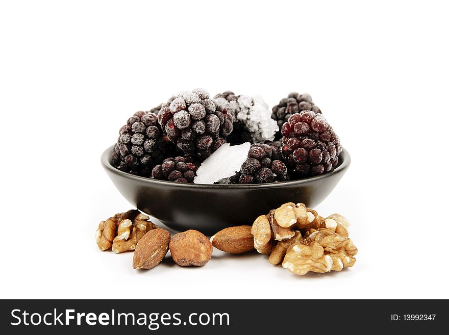 Ripe frozen blackberries in a small black bowl with mixed nuts on a reflective white background. Ripe frozen blackberries in a small black bowl with mixed nuts on a reflective white background