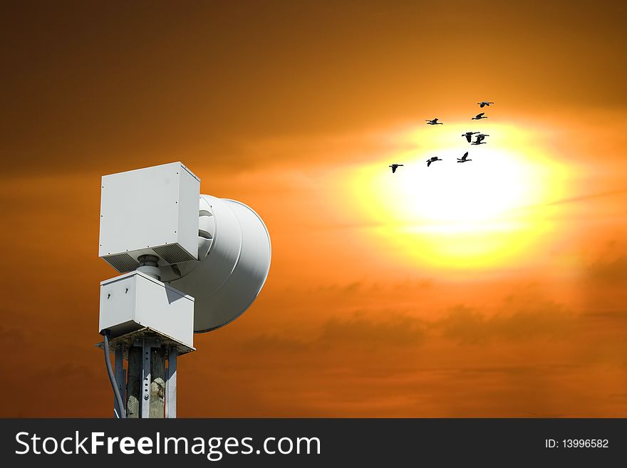 A community alert siren in the foreground with geese and sunset in the background. A community alert siren in the foreground with geese and sunset in the background.