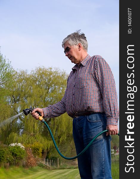 A mature man with grey hair and sunglasses using a hose to water the garden. A mature man with grey hair and sunglasses using a hose to water the garden.