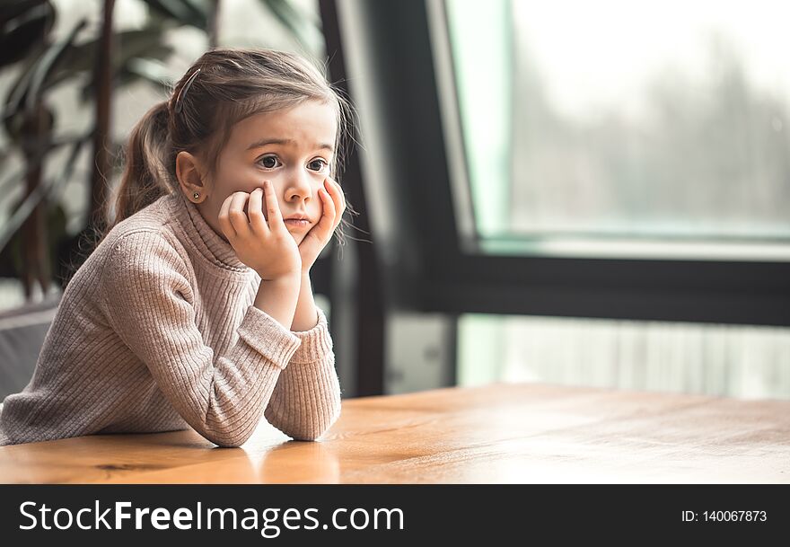 Charming little girl sitting at a wooden table by the window. Upset child