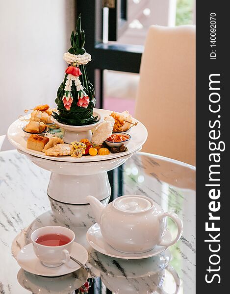 Thai assorted traditional dessert snack, curry puff, rice cracker, custard, spring rolls etc. on white ceramic pedestal tray with tea set. close up shot. Thai assorted traditional dessert snack, curry puff, rice cracker, custard, spring rolls etc. on white ceramic pedestal tray with tea set. close up shot