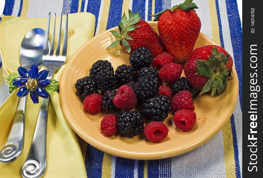 Assorted berries on a yellow plate on a striped textile with napkin, silverware, and napkin ring.