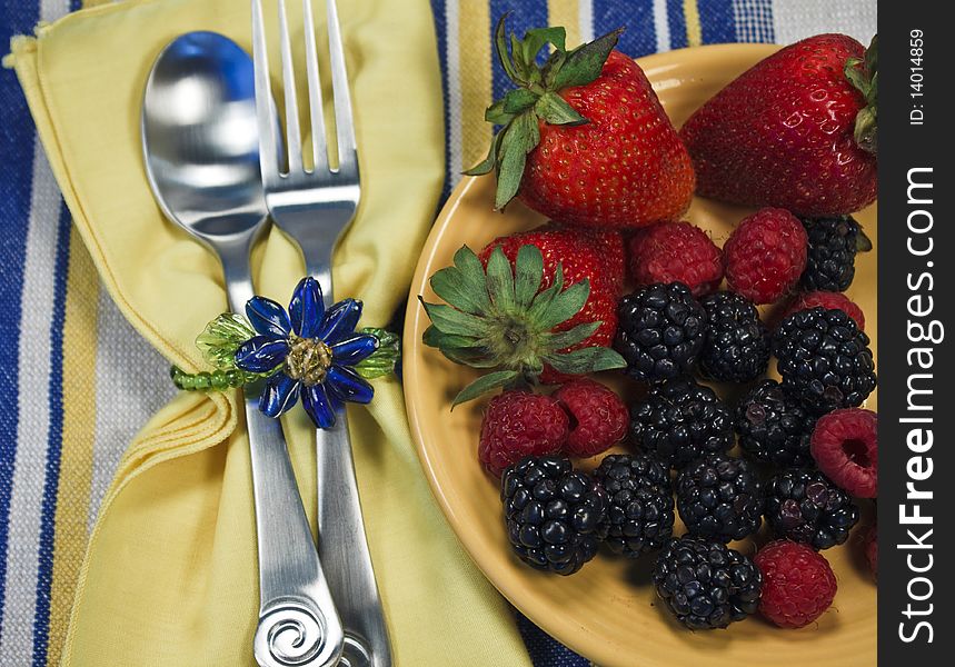 Assorted berries on a yellow plate on a striped textile with napkin, silverware, and napkin ring. Assorted berries on a yellow plate on a striped textile with napkin, silverware, and napkin ring.