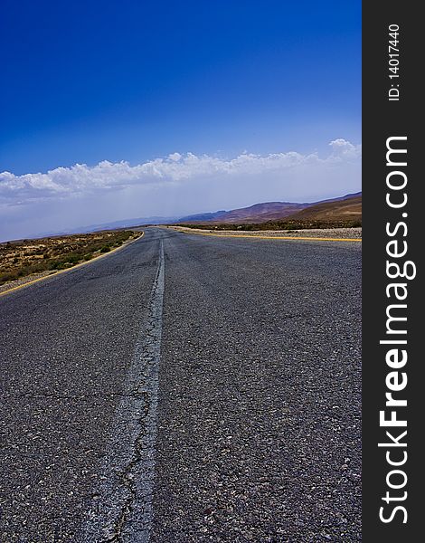 Photo of a long stretching Desert Road taken in a cloudy day, also showing a range of mountains at the horizon