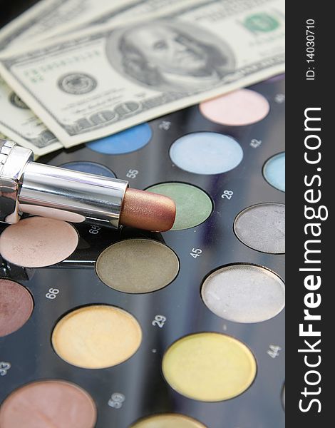 On a palette of powder brush and eye shadows there is lipstick in focus, on a background dollars. On a palette of powder brush and eye shadows there is lipstick in focus, on a background dollars