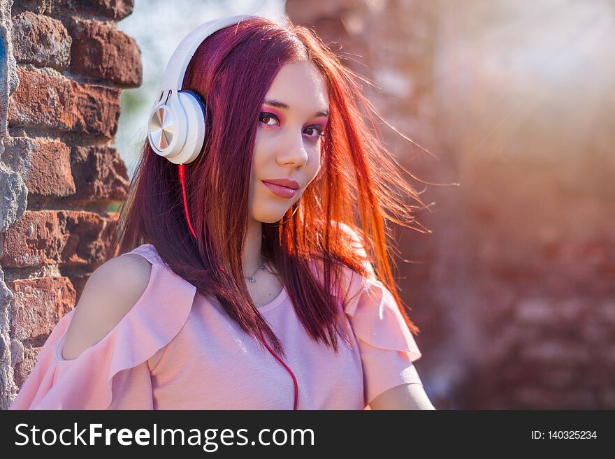 Gorgeous redheads lady listening music in headset on blurred background and red wall bricks in sunset. Charming girl with earphones enjoys with hair waving on wind. Close up, selective focus. Gorgeous redheads lady listening music in headset on blurred background and red wall bricks in sunset. Charming girl with earphones enjoys with hair waving on wind. Close up, selective focus