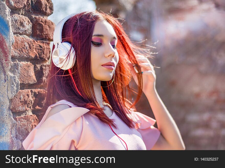 Gorgeous redheads lady listening music on headset and blurred outdoor on background and wall bricks. Charming girl with earphones enjoys with hair waving and eyes closed. Close up, selective focus. Gorgeous redheads lady listening music on headset and blurred outdoor on background and wall bricks. Charming girl with earphones enjoys with hair waving and eyes closed. Close up, selective focus