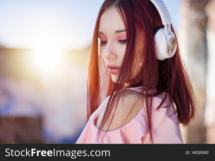 Gorgeous redhead lady listening music in headphones on blurred background outdoor