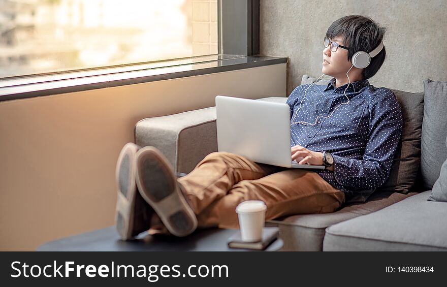 Young Asian man with headphones and glasses sitting on sofa looking out of window while watching movie from laptop computer. Urban lifestyle in living room. Relaxation concept. Young Asian man with headphones and glasses sitting on sofa looking out of window while watching movie from laptop computer. Urban lifestyle in living room. Relaxation concept